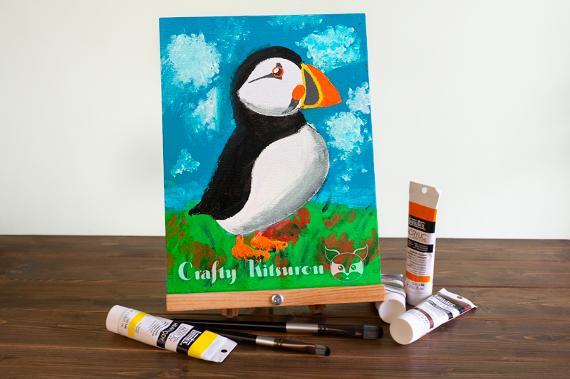 Acrylic painting of a puffin surrounded by tubes of paint and brushes on a dark wood surface.