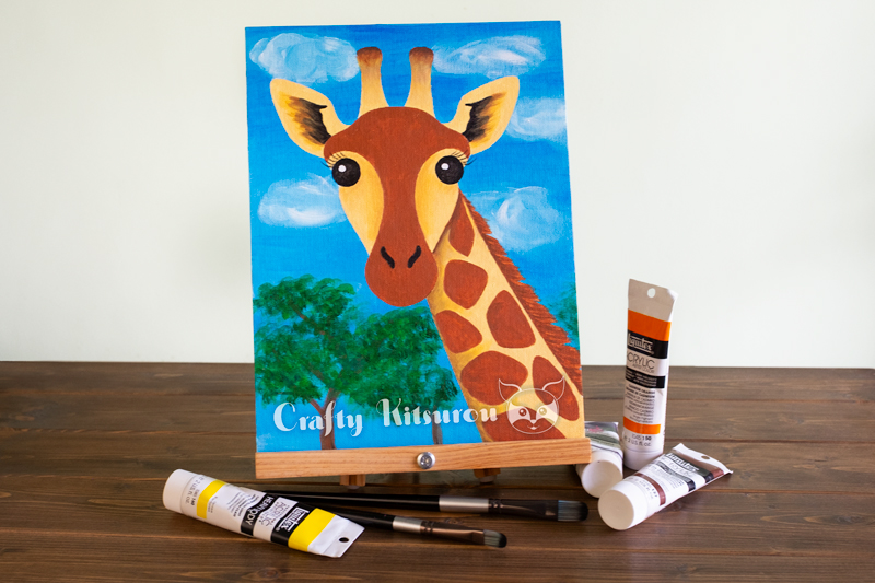 Acrylic painting of a giraffe surrounded by tubes of paint and brushes on a dark wood surface.